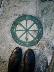 Pair of black shoes at bottom of picture stand next to compass embedded in rock