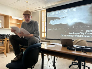 Prof. Sean Holman of the Climate Disaster Project sits on a desk in front of projection screen reading from a paper document.